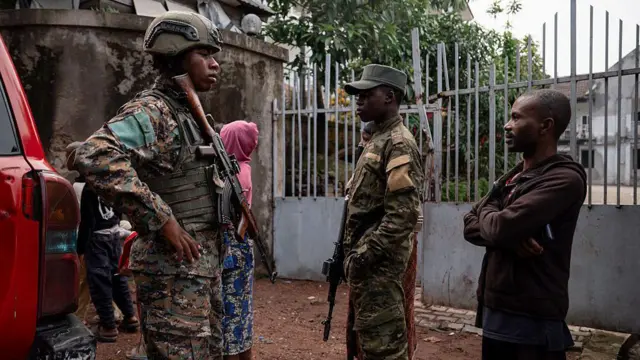 Deux militaires en tenue de combat accompagnés d'un civil en sweat-shirt marron et d'une femme en sweat-shirt rose, dans une zone de conflit